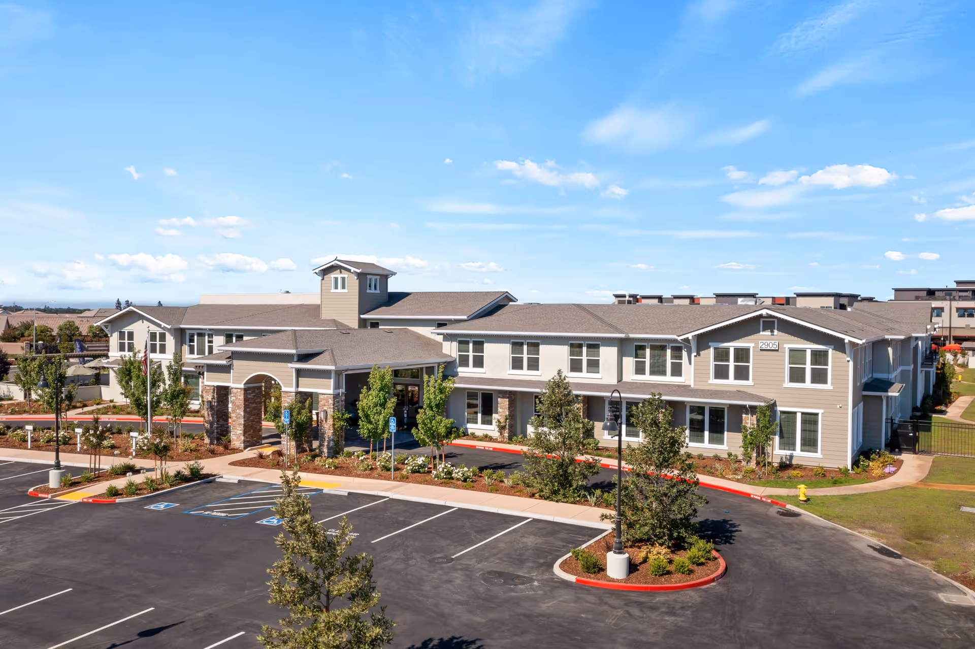 Exterior view of Oakmont of Lodi senior living facility showing a large two-story building with beige siding and stone accents, surrounded by landscaped greenery and a mostly empty parking lot under a bright blue sky with scattered clouds.