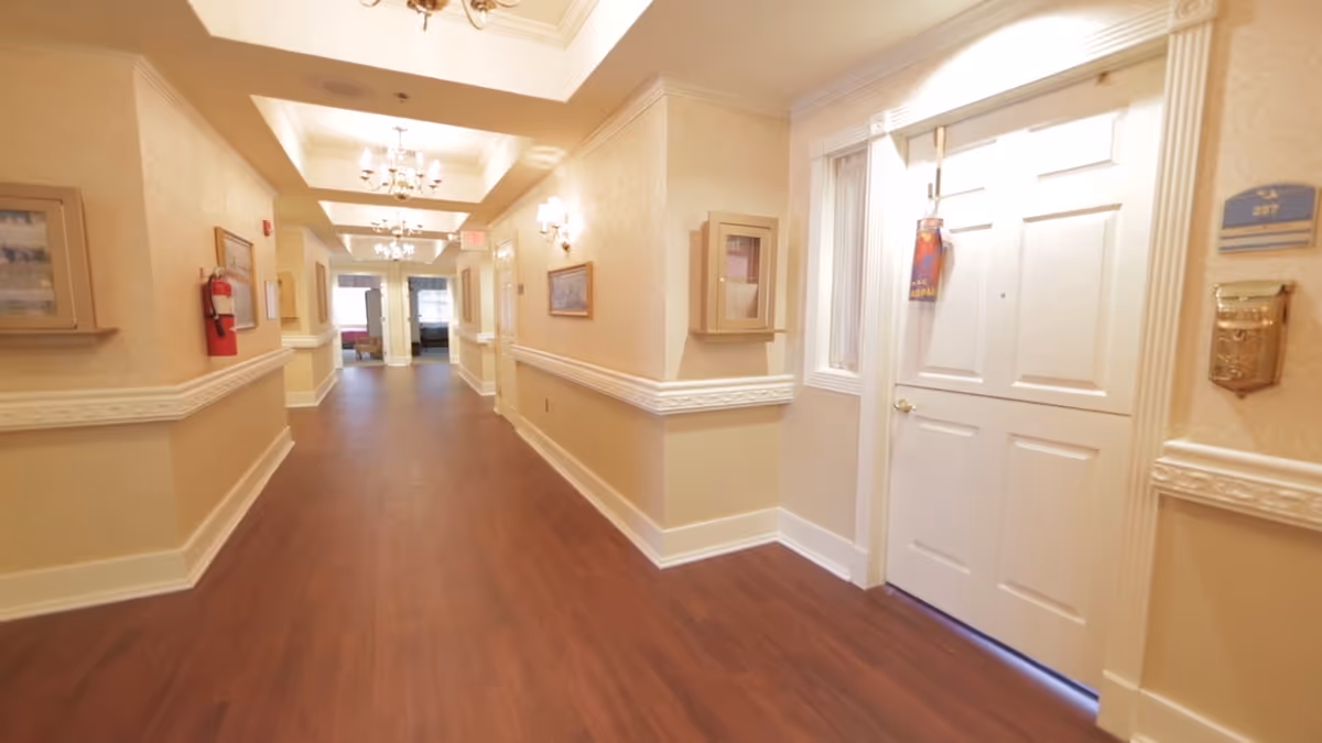 A well-lit hallway in an assisted living facility with wooden flooring, beige walls with white trim, chandeliers on the ceiling, framed pictures on the walls, and a white door labeled 227 on the right side.