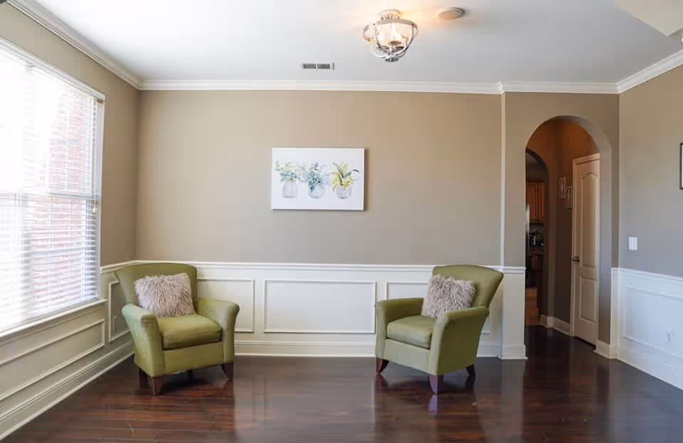 A bright living room with two green armchairs each with a fluffy pillow, positioned on a dark wooden floor. The walls are beige with white wainscoting, and a painting of three potted plants hangs on the wall. There is a large window with blinds on the left side and an arched doorway leading to another room on the right.
