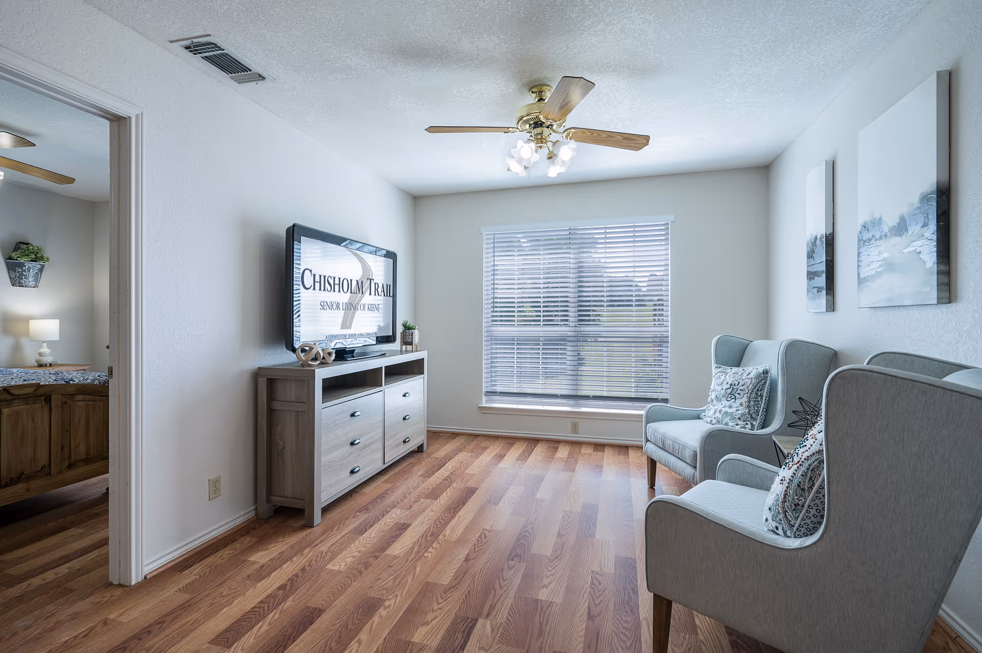 A bright living room with wooden flooring, two light gray armchairs with patterned cushions, a wooden TV stand with a flat-screen TV displaying 'Chisholm Trail Senior Living of Denton', and a large window with blinds letting in natural light. The room has white walls, two framed artworks, and a ceiling fan with lights. A doorway on the left leads to a bedroom with a wooden bed and a lamp.