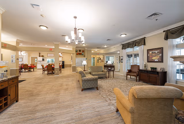 A spacious and well-lit common area in a senior living facility featuring comfortable armchairs and sofas arranged on a patterned carpet. In the background, there are dining tables and chairs, large windows with curtains, framed artwork on the walls, and warm ceiling lights. The area has a welcoming and cozy atmosphere.