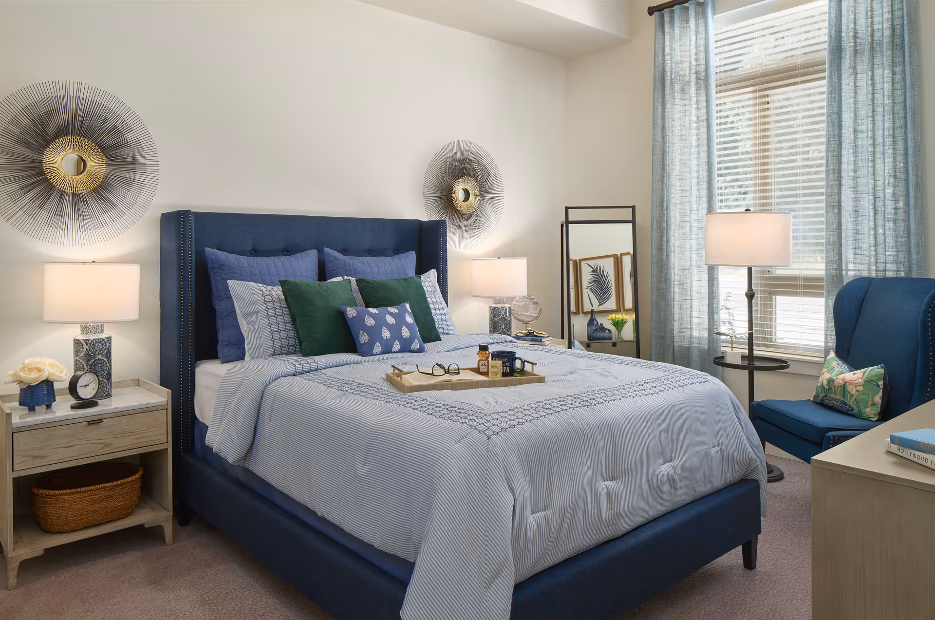 A well-decorated bedroom with a blue upholstered bed frame and a neatly made bed with blue and green pillows. There are two bedside tables with lamps, a small clock, and a flower vase. A full-length mirror stands near the window with light blue curtains, and a blue armchair with a decorative pillow is placed next to a floor lamp. The room has a calm and cozy ambiance with soft natural light coming through the window.