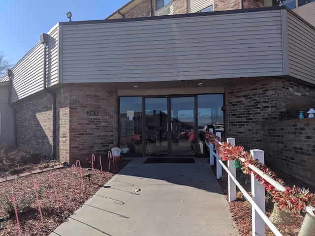 Entrance to a brick building with glass double doors and a concrete walkway. The walkway is decorated with candy cane stakes and garlands on the white railing. The building number 2801 is visible on the left side of the entrance.