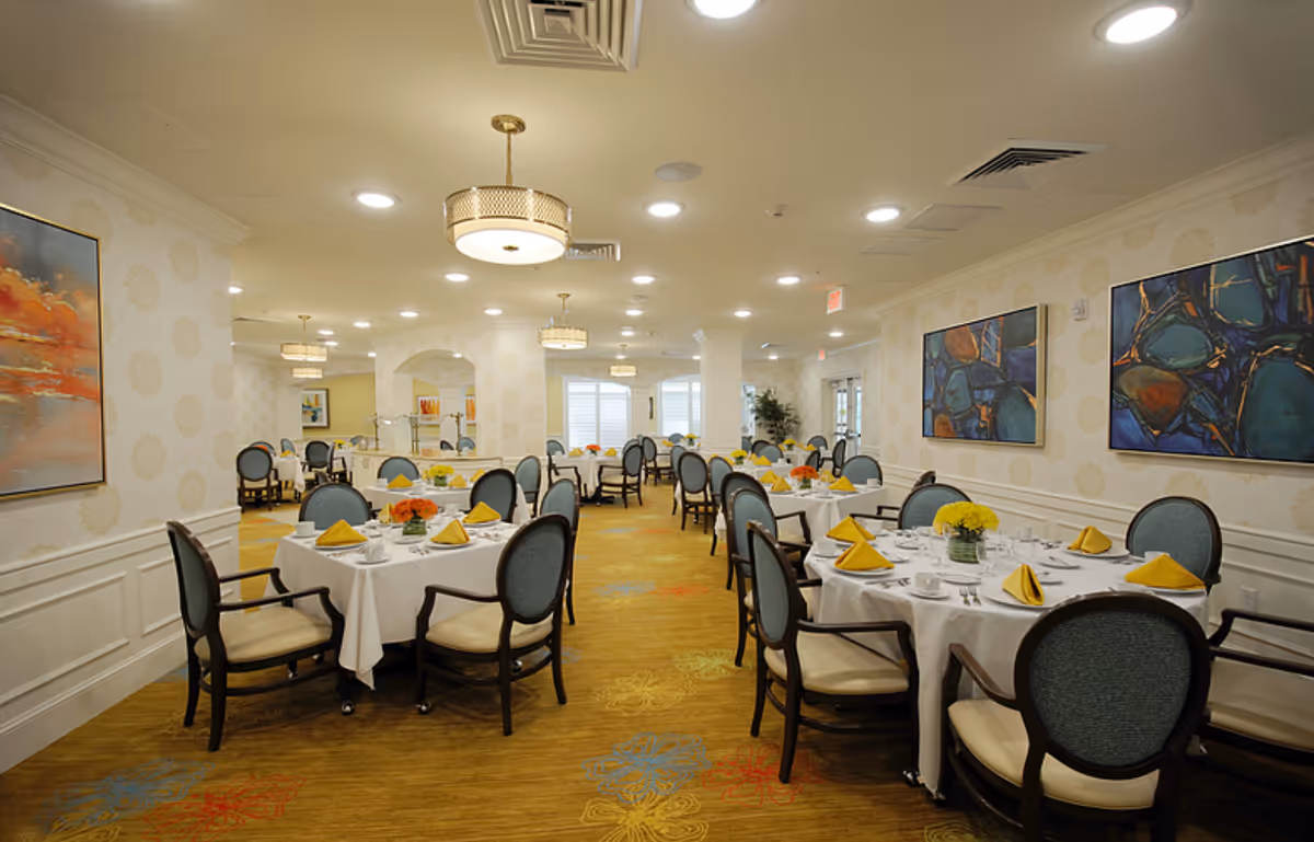 A spacious dining room with multiple round tables covered with white tablecloths, each set with yellow folded napkins, white cups, and silverware. The chairs have dark wooden frames with light blue upholstered backs and cream-colored seats. The room is well-lit with ceiling lights and decorative hanging lamps. The walls are decorated with patterned wallpaper and colorful abstract paintings. The carpet has a floral pattern in various colors.