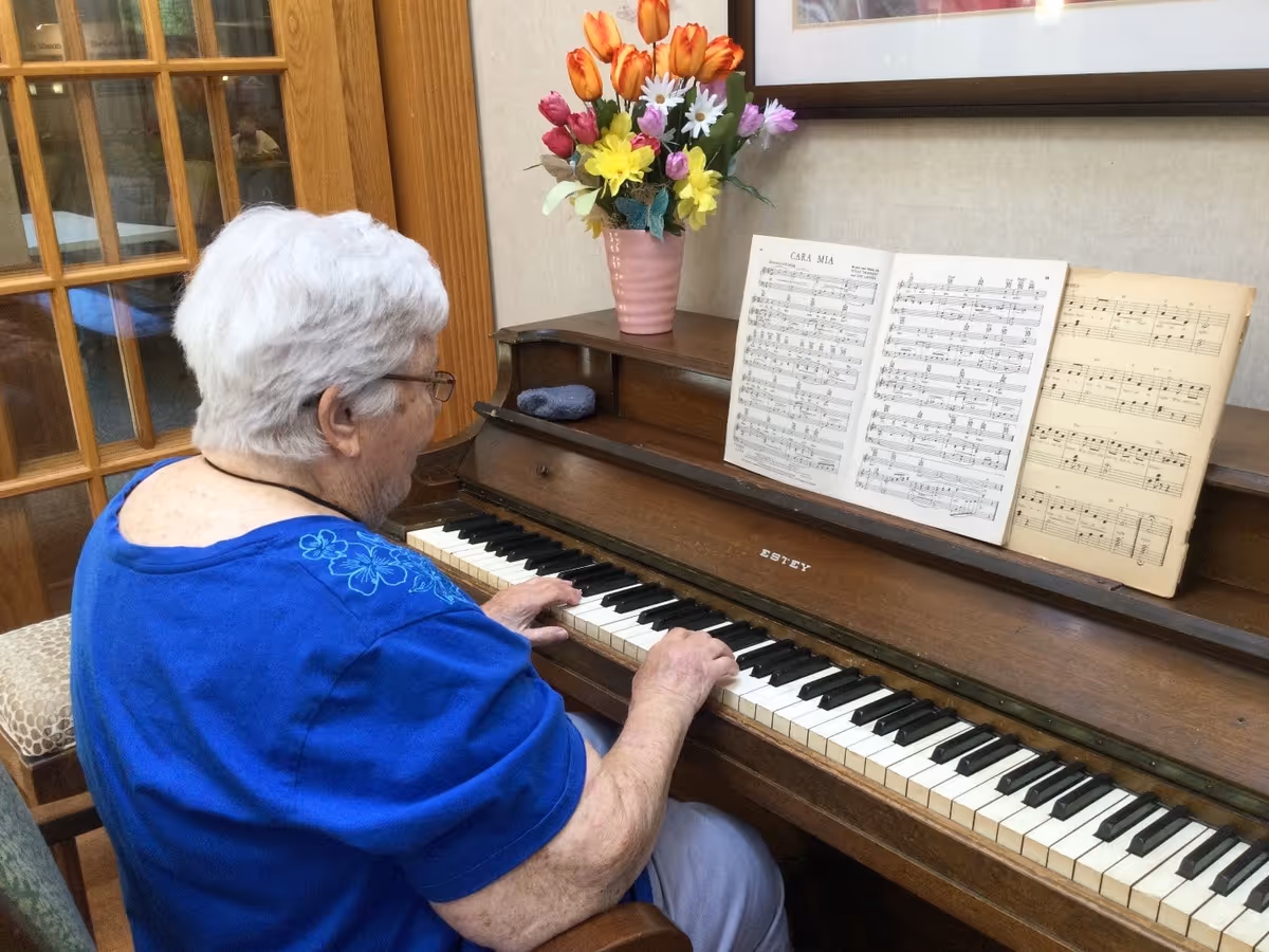 An elderly woman with white hair and glasses is playing a wooden piano with sheet music titled 'CARA MIA' on the music stand. A vase with colorful artificial flowers is placed on top of the piano. The setting appears to be a cozy indoor room with wooden furniture and a glass-paneled door in the background.