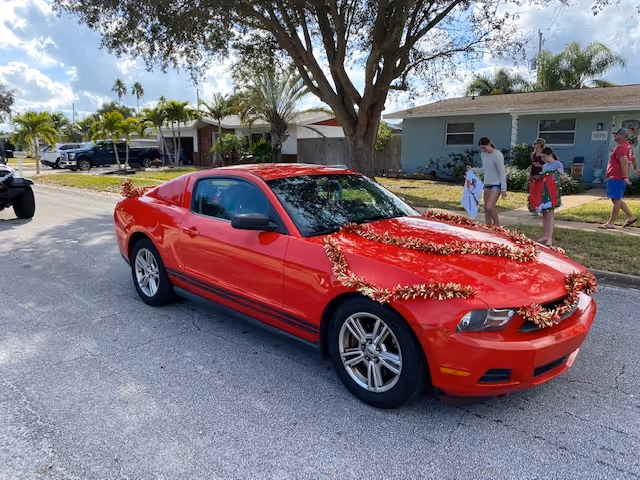 A red sports car decorated with tinsel garlands parked on a residential street with houses and people walking on the sidewalk.