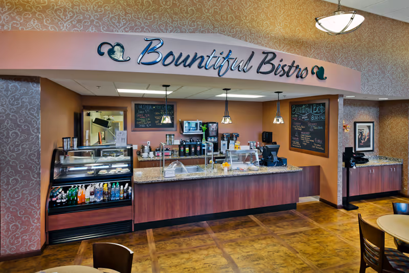 Interior view of Bountiful Bistro at Grand Arbor by Vivie, featuring a counter with a display case of beverages and snacks, coffee machines, and menu boards on the wall. The area has warm-toned walls and patterned wallpaper with hanging pendant lights above the counter.