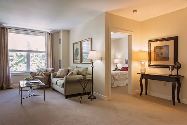 A bright living room with a large window covered by curtains, a green sofa with cushions, an armchair, a glass coffee table, and a floor lamp. To the right, there is an open doorway leading to a bedroom with a bed and bedside lamps. A console table with a lamp and framed artwork is against the wall near the doorway.