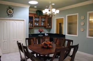 Interior dining area with a round wooden table surrounded by six chairs. A chandelier hangs above the table, which has a floral centerpiece. Behind the table is a built-in cabinet with glass doors displaying dishes and decorative items. The walls are painted green with white trim, and there are two windows and three decorative plates on one wall. A clock is mounted above a set of double doors.