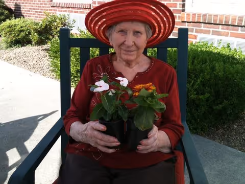An elderly woman wearing a red wide-brimmed hat and a red top is sitting outdoors on a green chair, holding two small potted flowering plants. There is a brick wall and green bushes in the background.