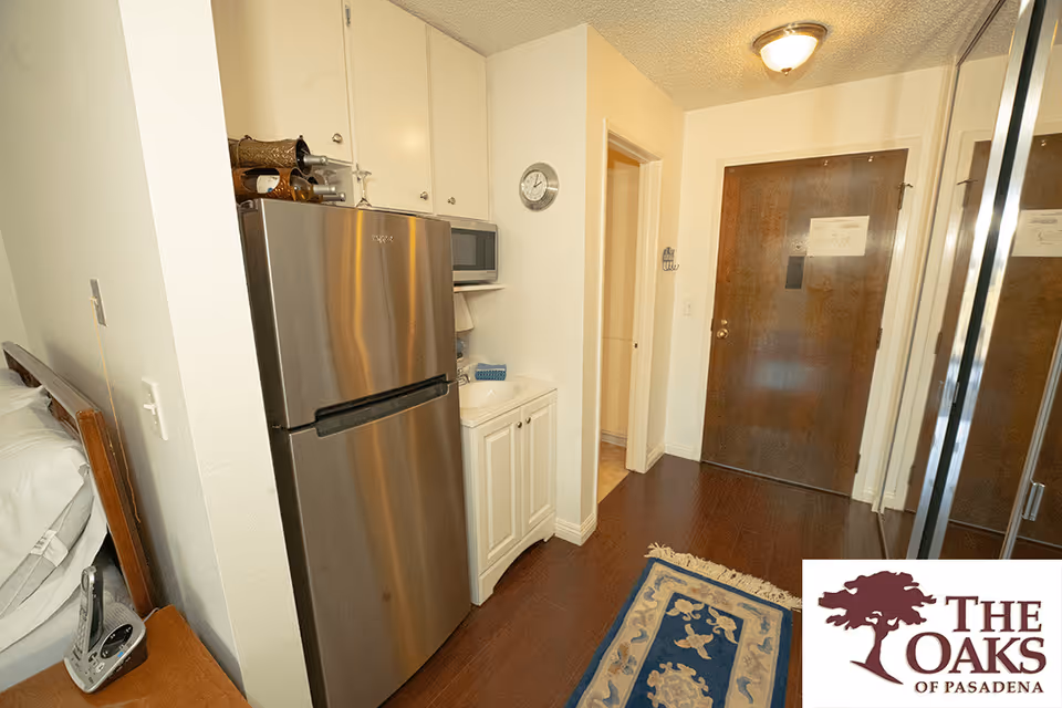Interior view of a small apartment entryway with a wooden front door, a stainless steel refrigerator, a microwave on a shelf above a white cabinet, a wall clock, a blue and beige patterned rug on wooden flooring, and a mirrored closet door on the right. Part of a bed and a nightstand with a phone are visible on the left.