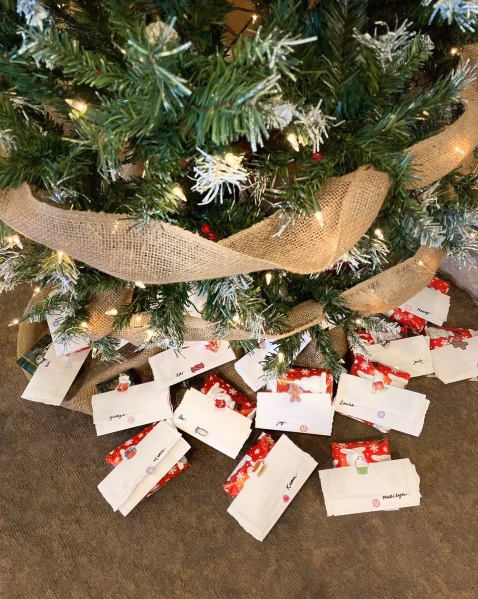 Close-up view of the base of a decorated Christmas tree with burlap ribbon and white lights. Underneath the tree are multiple wrapped gifts and white envelopes with handwritten names and festive stickers on them, placed on a carpeted floor.