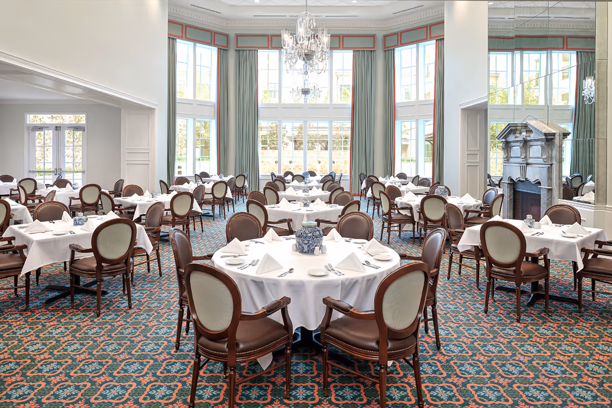 A spacious, elegant dining room with multiple round and rectangular tables covered with white tablecloths and set with napkins, plates, and silverware. The room features large windows with green curtains, a decorative chandelier hanging from the ceiling, and a fireplace with a mirror above it. The carpet has a patterned design in green and red tones.