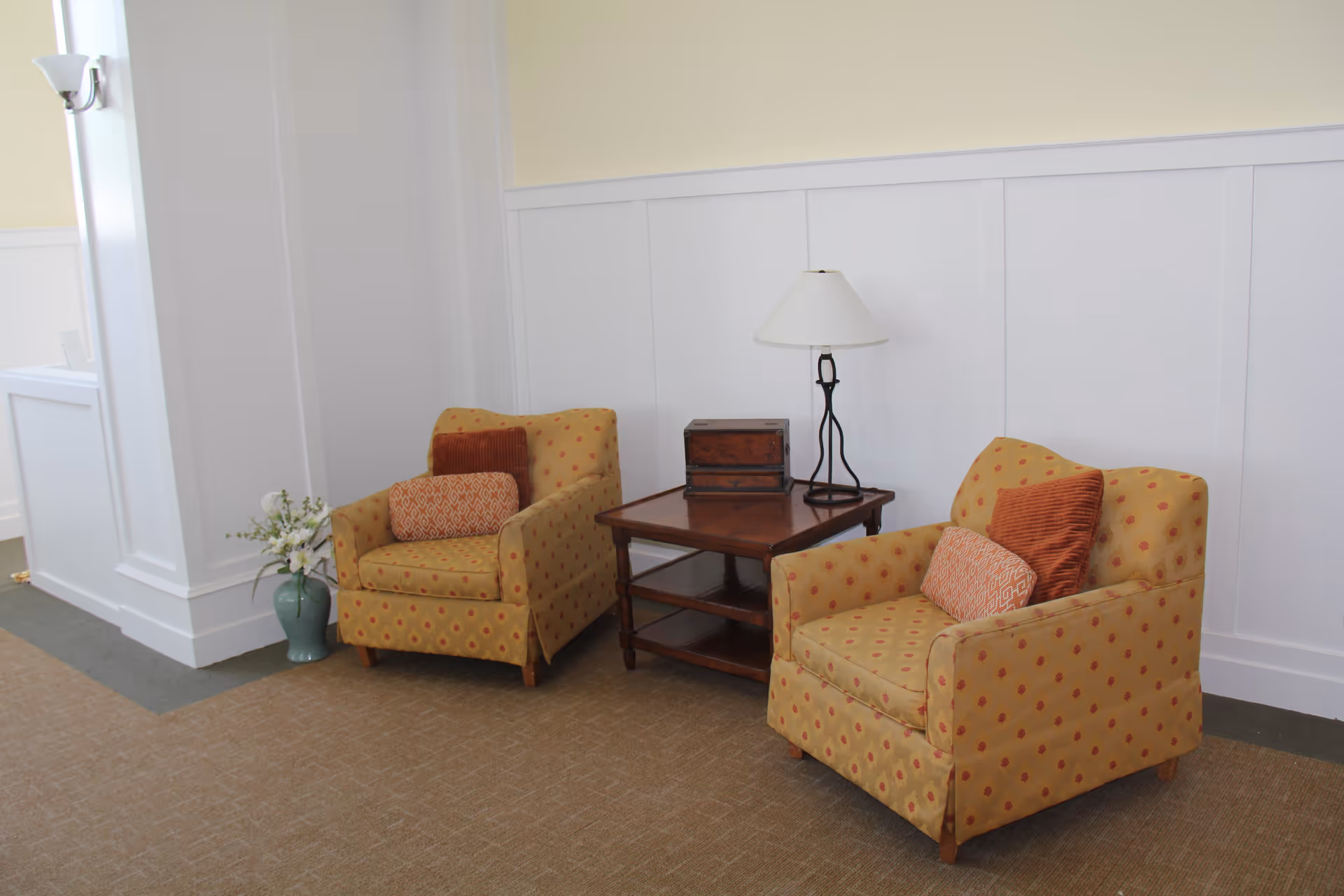 Two patterned armchairs with orange and red cushions are placed on either side of a wooden side table with a lamp and a decorative box. A green vase with white flowers is on the floor next to one chair. The setting is indoors with white paneled walls and beige carpet.
