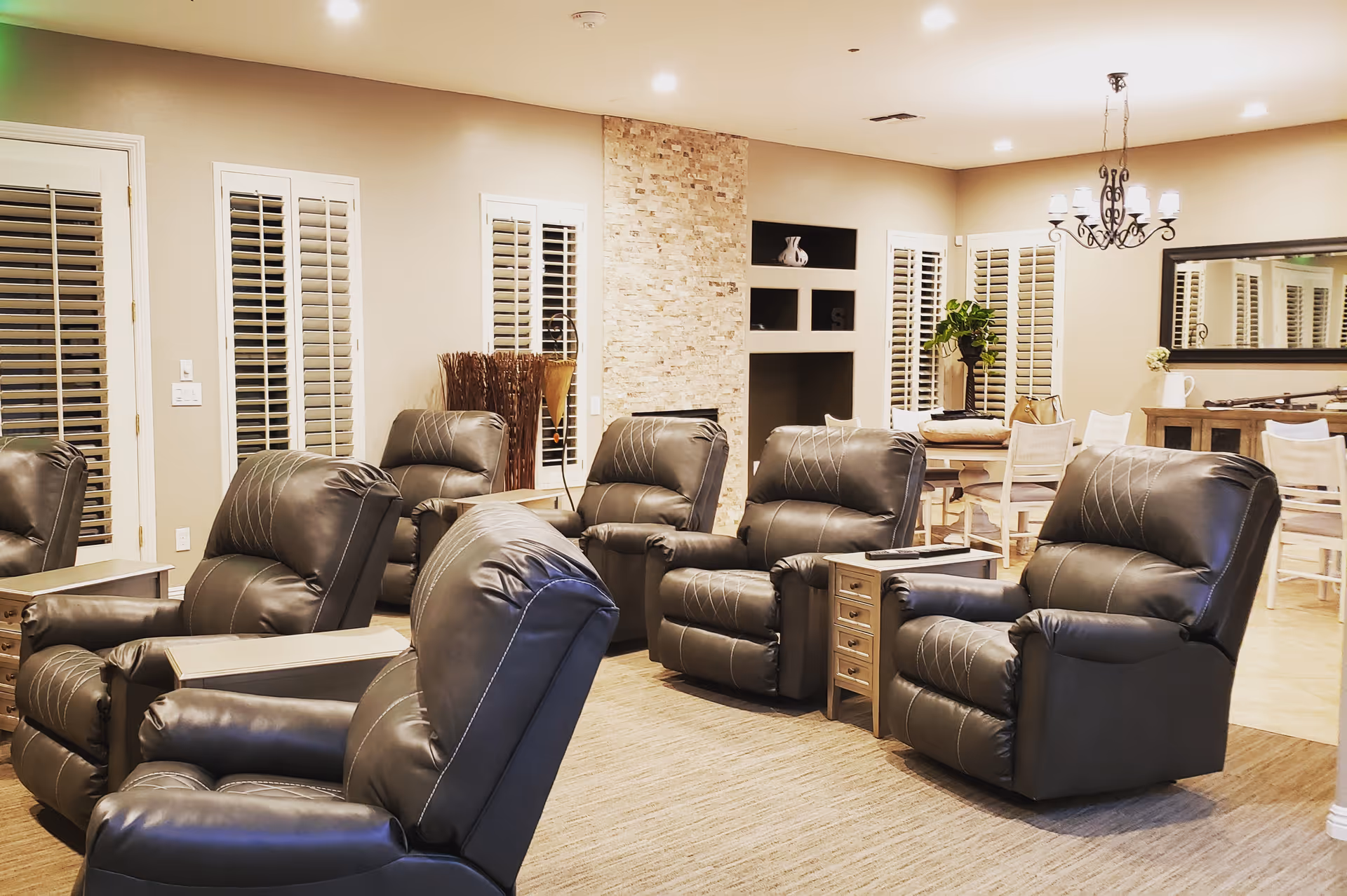A cozy senior living facility common area with several black leather recliners arranged in a semi-circle facing a stone accent wall with built-in shelves. The room has beige walls, white plantation shutters on the windows, a chandelier, a large mirror on the wall, and a dining table with chairs in the background.