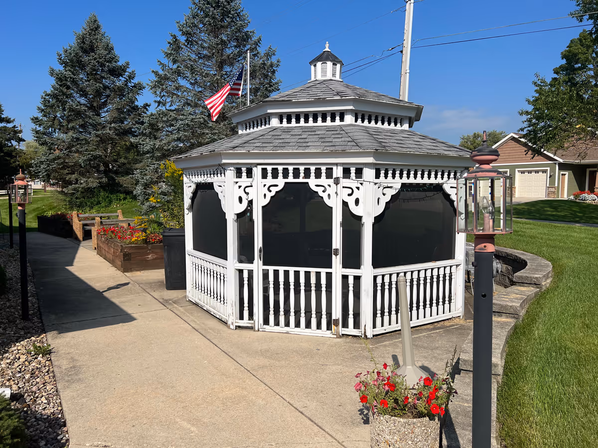 A white decorative gazebo sits on a paved walkway in a landscaped outdoor courtyard with flower planters and an American flag.