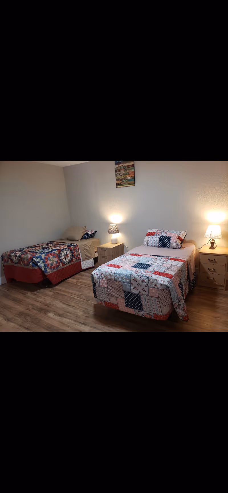 A bedroom with two single beds, each with colorful patchwork quilts. There are two wooden nightstands between and beside the beds, each with a lit table lamp. The room has wooden flooring and plain light-colored walls with a small decorative wall hanging above the nightstands.