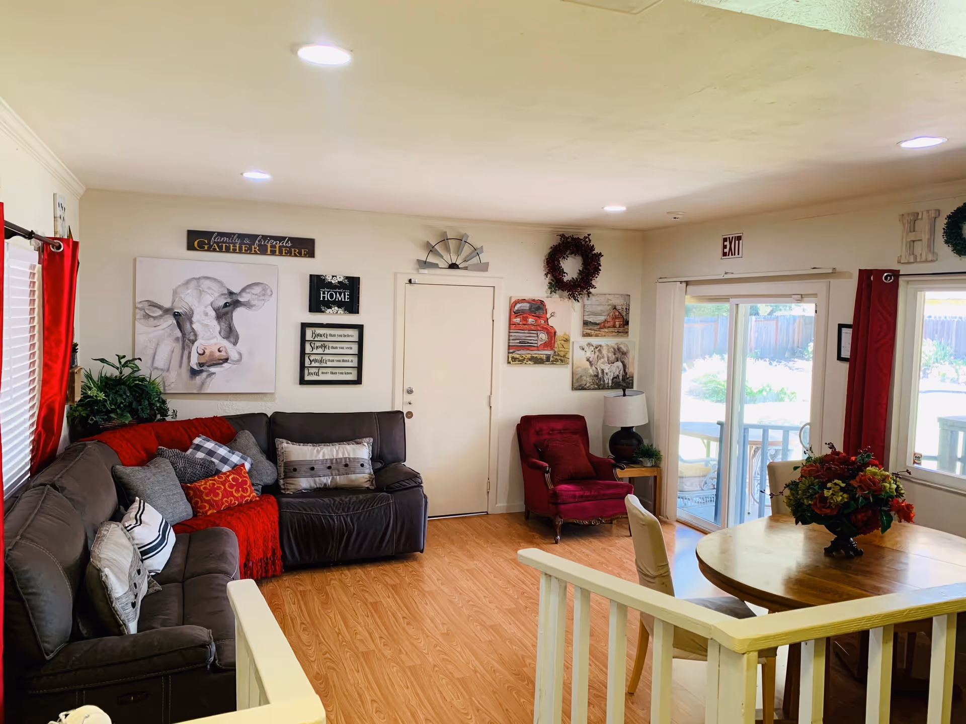 A cozy living room with a dark brown sectional sofa adorned with various pillows and a red throw blanket. There is a red armchair next to a small side table with a lamp. The walls are decorated with paintings, including a cow, a red truck, and a sheep, along with decorative signs and wreaths. A round wooden dining table with chairs and a floral centerpiece is visible near sliding glass doors that lead outside. The room has wood flooring and recessed ceiling lights.