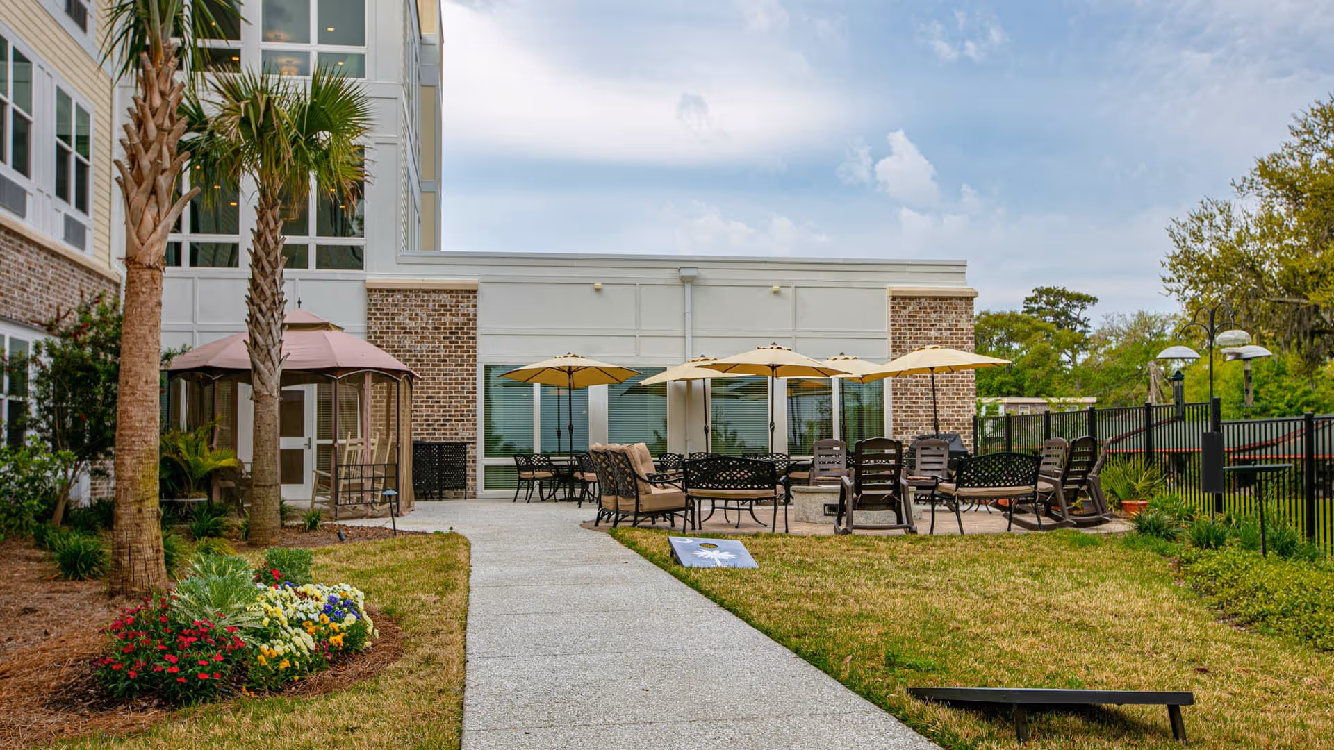 Outdoor patio area at The Retreat at Lady's Island featuring a paved walkway, palm trees, flower beds, multiple seating arrangements with chairs and tables under beige umbrellas, and a screened gazebo attached to the building.