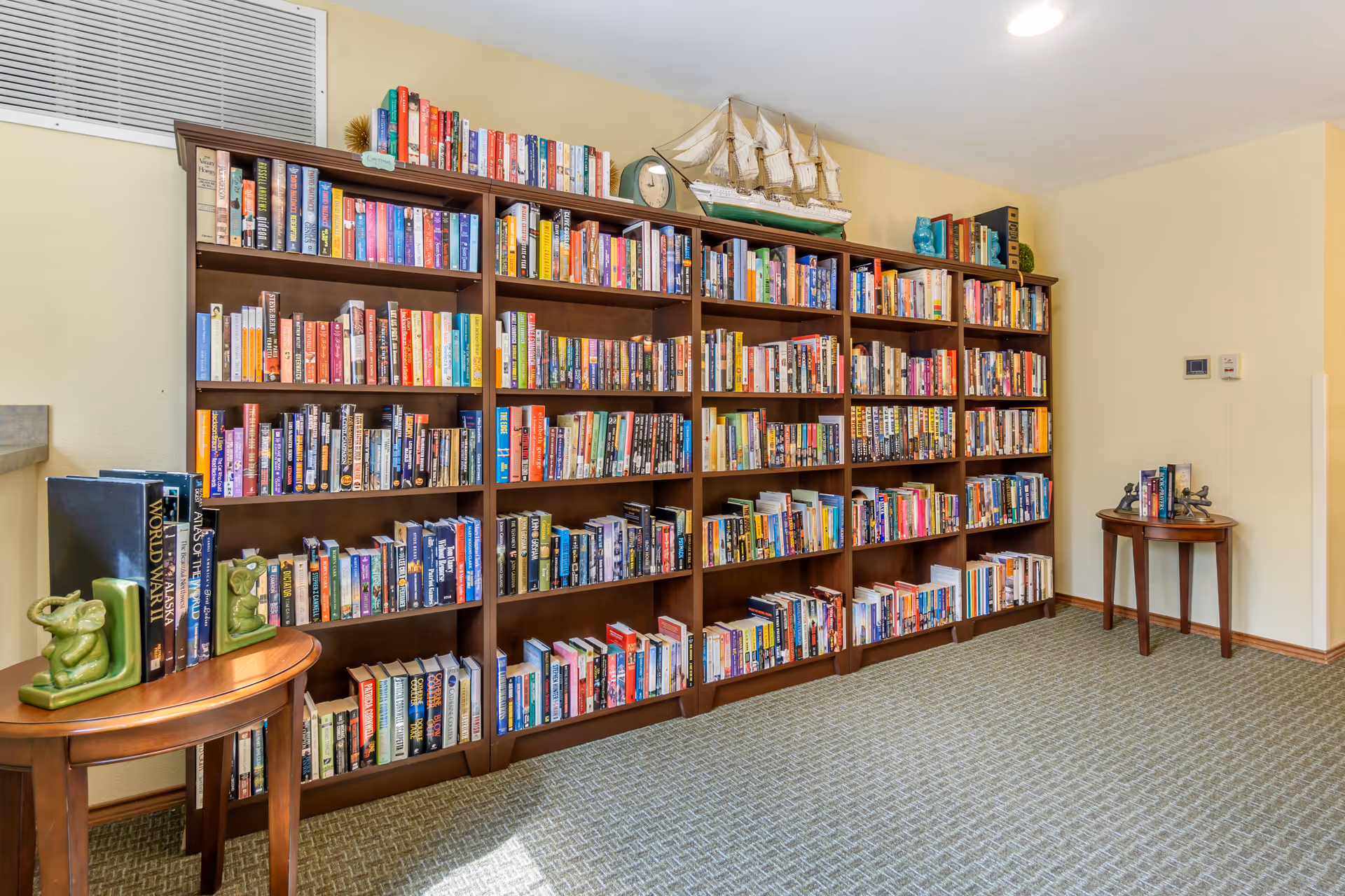 A wall of wooden bookshelves filled with books and decorative items in a well-lit room with two small round tables.