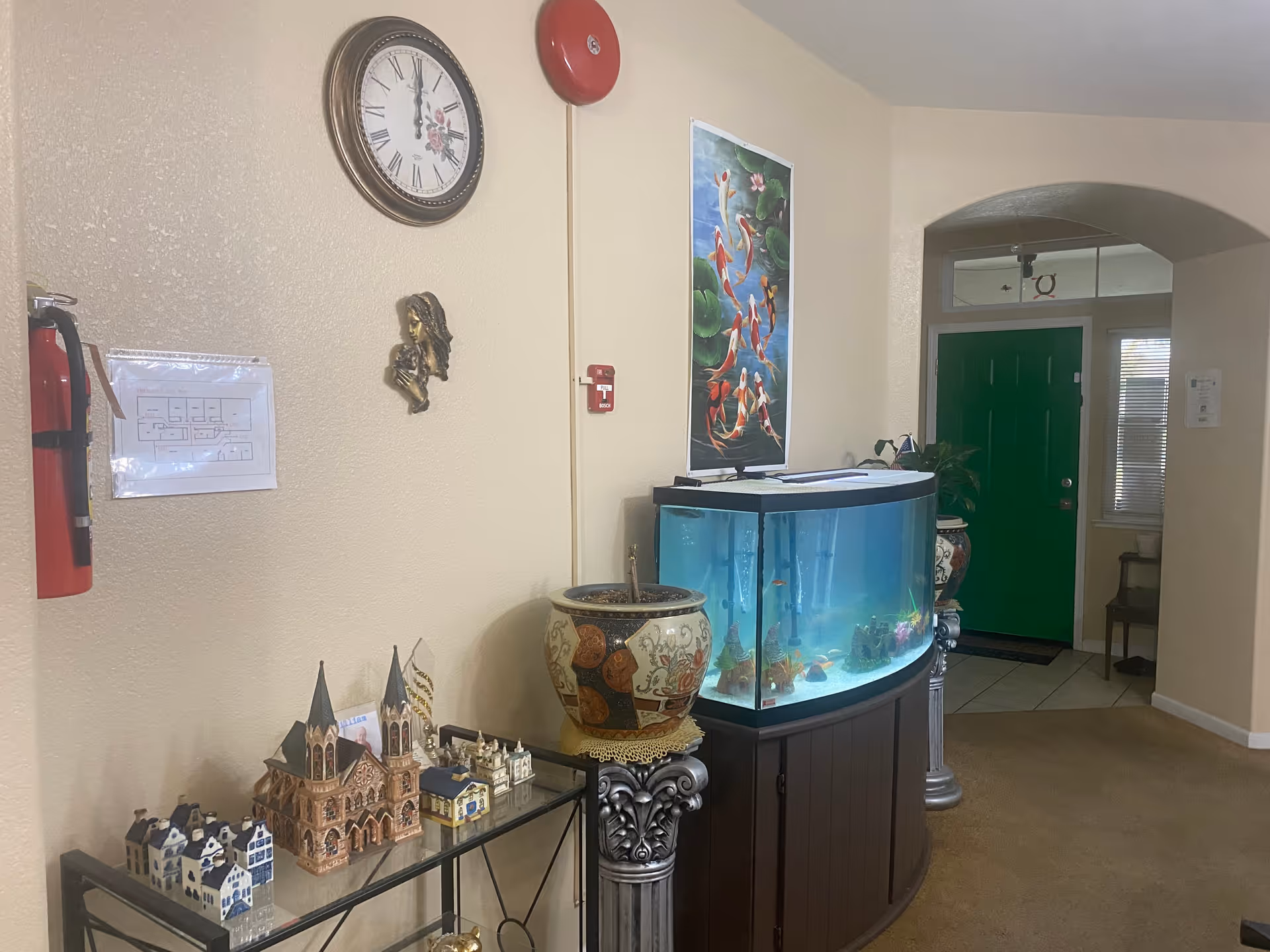 Interior entry area with a curved aquarium on a wooden stand, miniature building models on a glass table, wall clock, and a green front door.