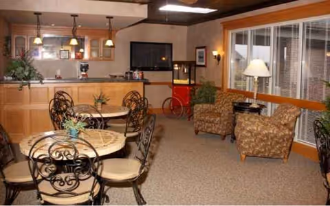 Indoor senior living common area with round dining tables, chairs, and a seating nook near windows.