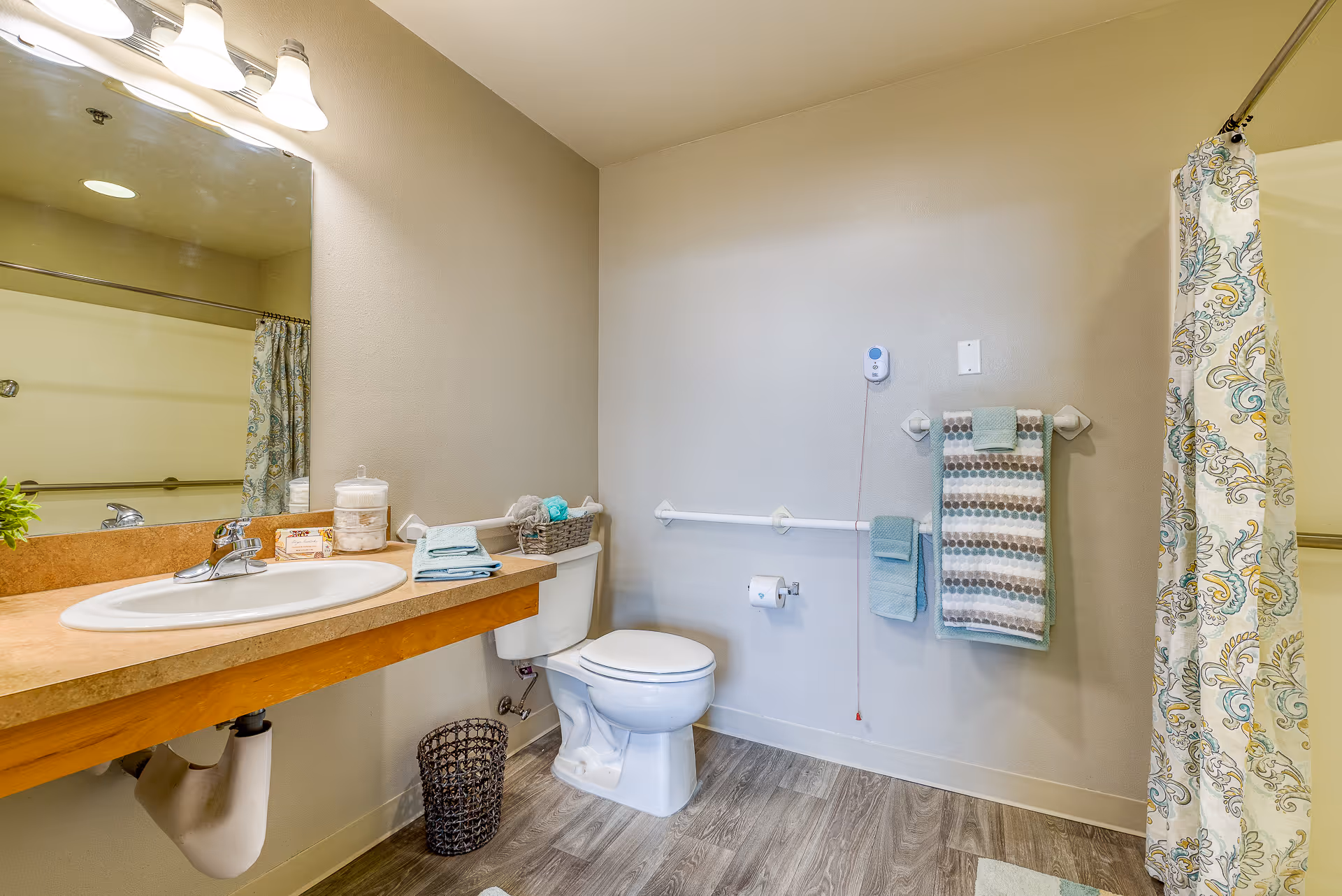 A clean and well-lit bathroom featuring a sink with a countertop, a large mirror, a toilet with grab bars on the wall, a wastebasket, and a shower area with a patterned shower curtain. Towels are neatly hung on the wall, and a basket with toiletries is placed on the toilet tank.