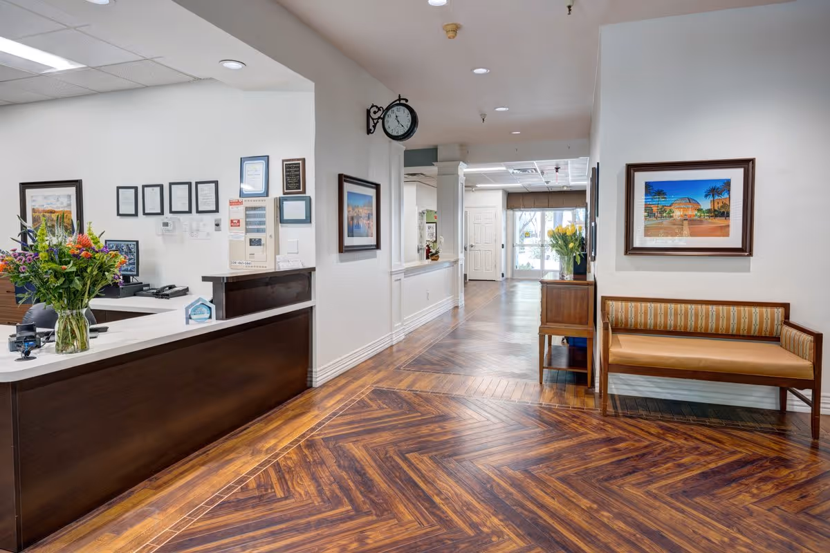 Reception area of The Oaks at Inglewood facility featuring a wooden front desk with a vase of colorful flowers, a wall clock, framed pictures on white walls, a wooden bench with striped cushions, and a hallway with wooden flooring leading to glass doors at the far end.