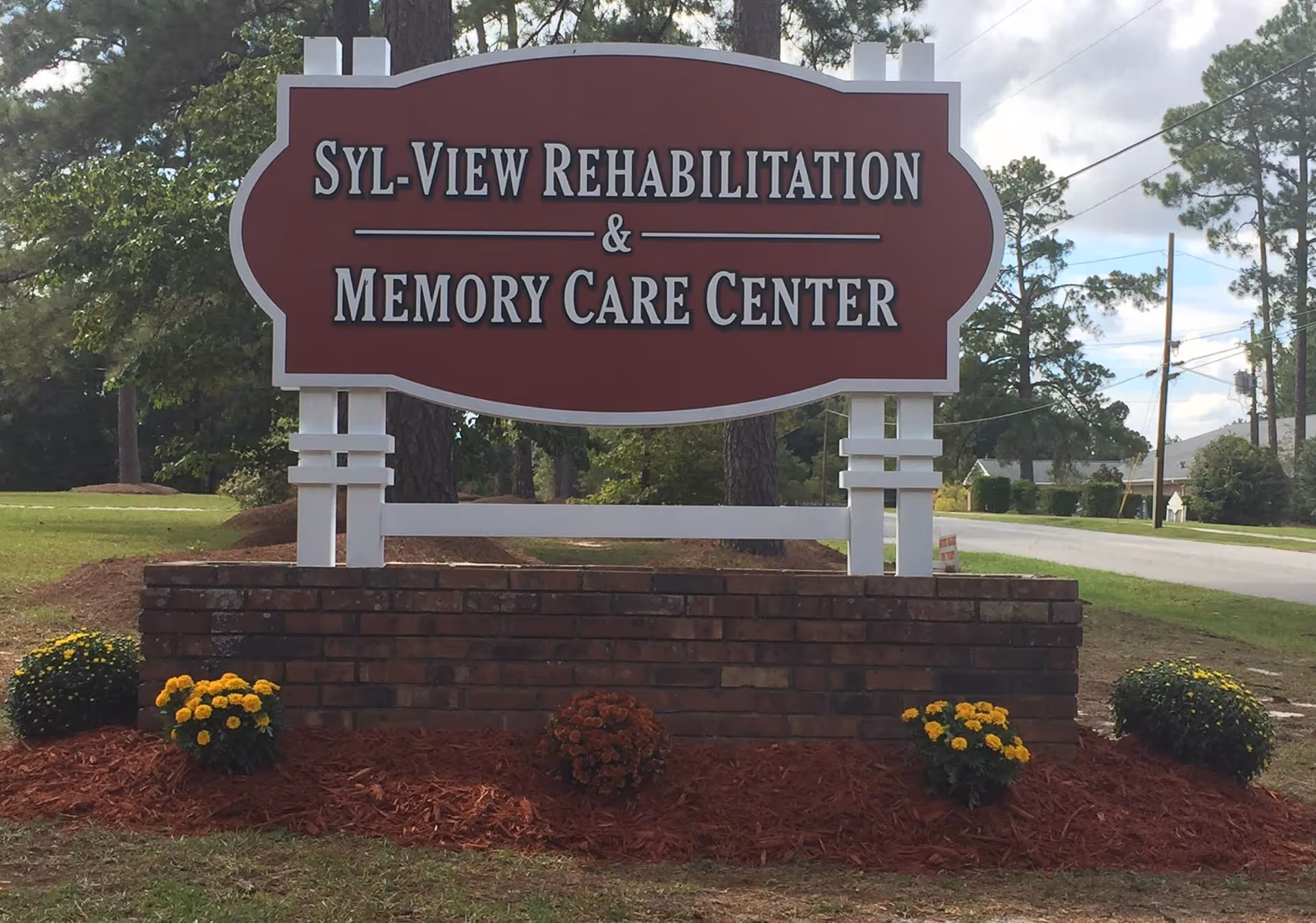 A large maroon and white sign reading 'Syl-View Rehabilitation & Memory Care Center' on a brick base with flowers and trees in the background.