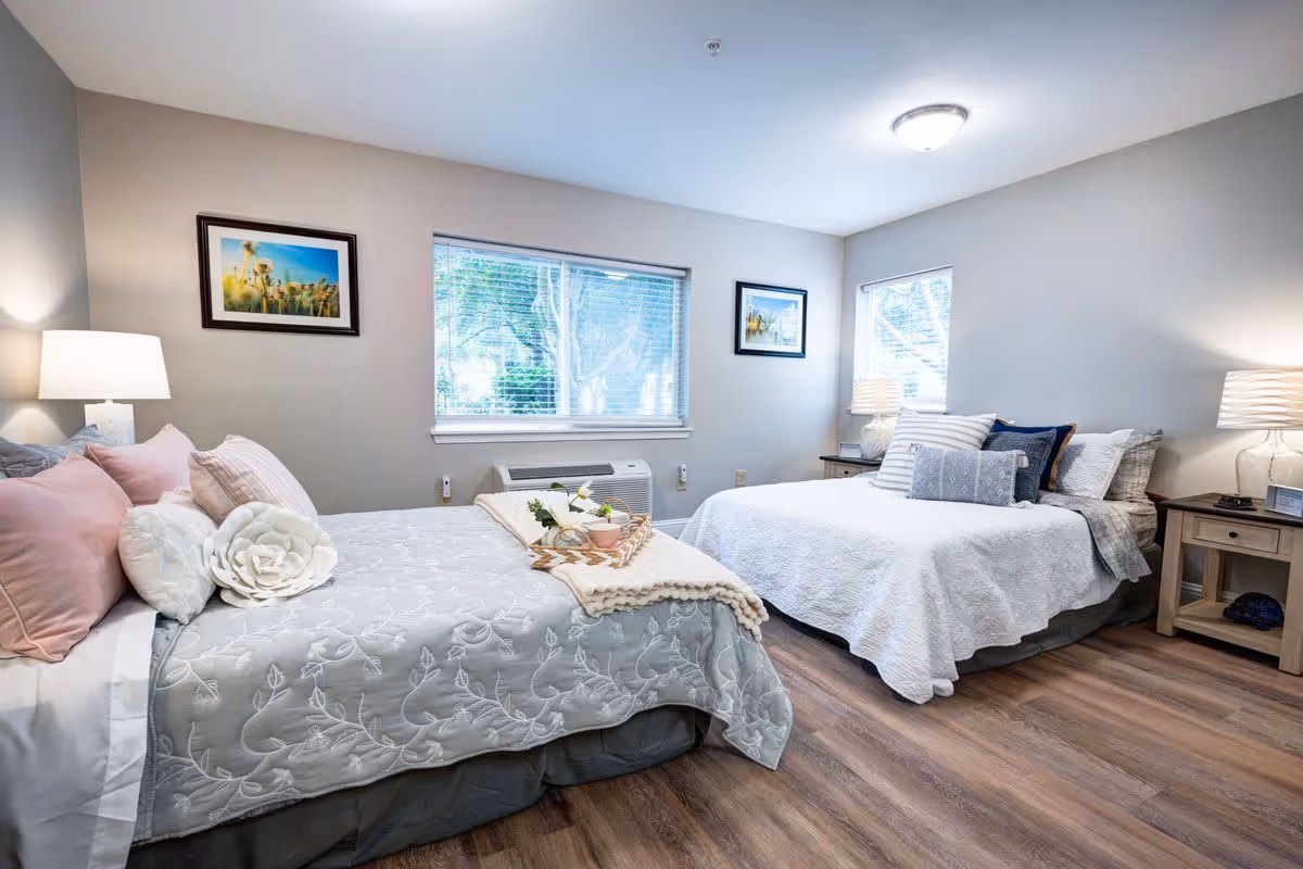 A bright and cozy bedroom with two beds, each adorned with multiple pillows and soft bedding. The room features two windows with blinds, two bedside tables with lamps, and framed artwork on the walls. The floor is wooden, and the overall decor is light and inviting.