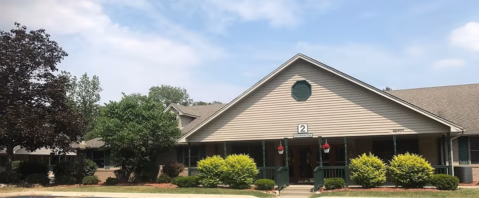 Exterior view of a single-story building with beige siding and a peaked roof under a partly cloudy sky. The building has a covered porch with green railings and hanging flower baskets. There are several bushes and trees in front of the building, and a sign above the entrance reads '2 INFORMATION'.
