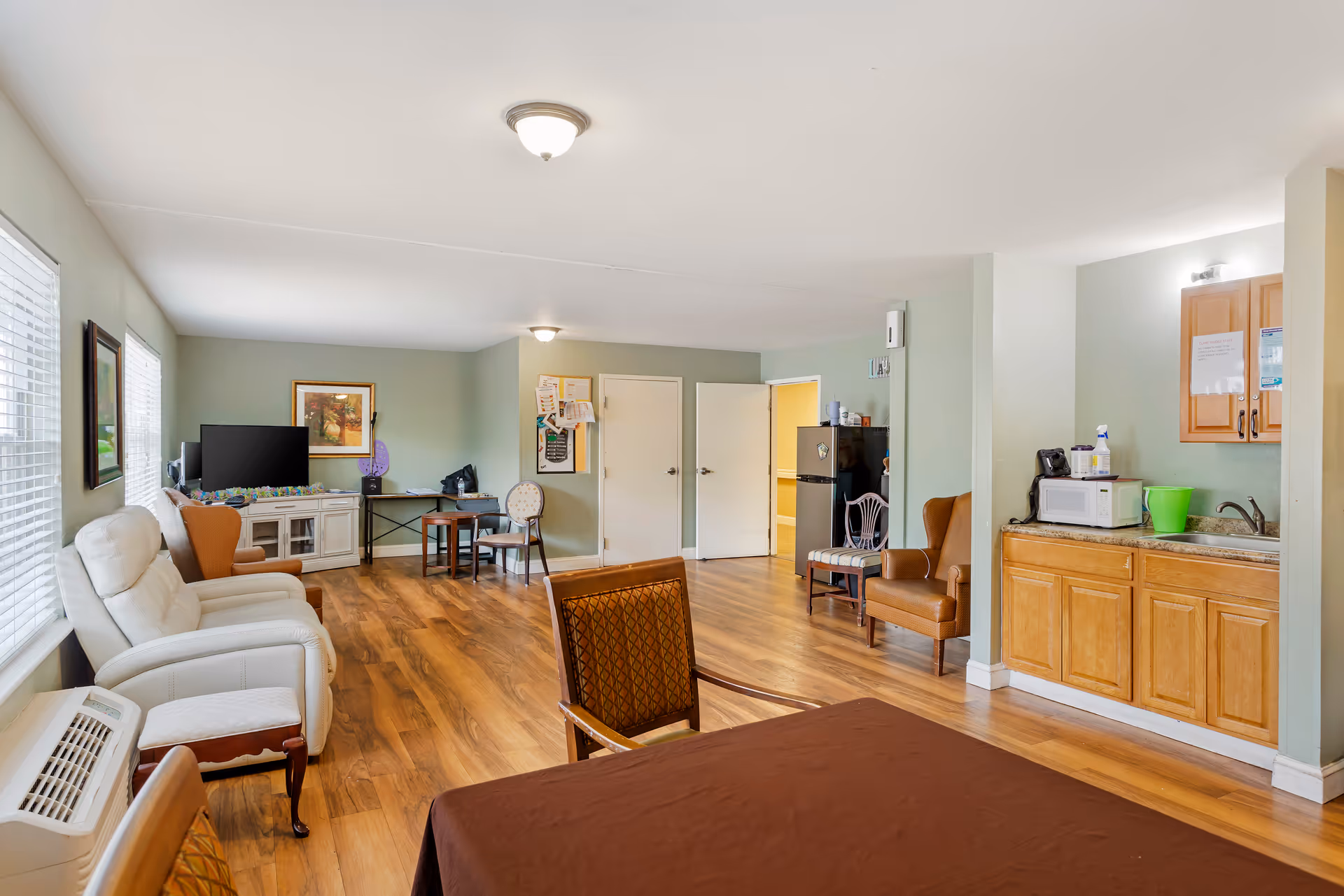 A spacious common area in a senior living facility with wooden flooring and light green walls. The room features multiple seating options including white and brown armchairs, a small table with chairs, a TV on a white stand, and a kitchenette area with a microwave, sink, and cabinets. There are two doors in the background and windows on the left side letting in natural light.