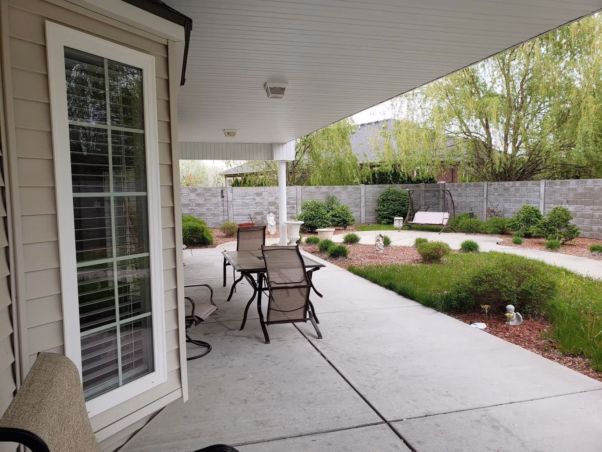 Covered patio with a table and chairs overlooking a landscaped courtyard with shrubs, a walkway, and a swing bench.