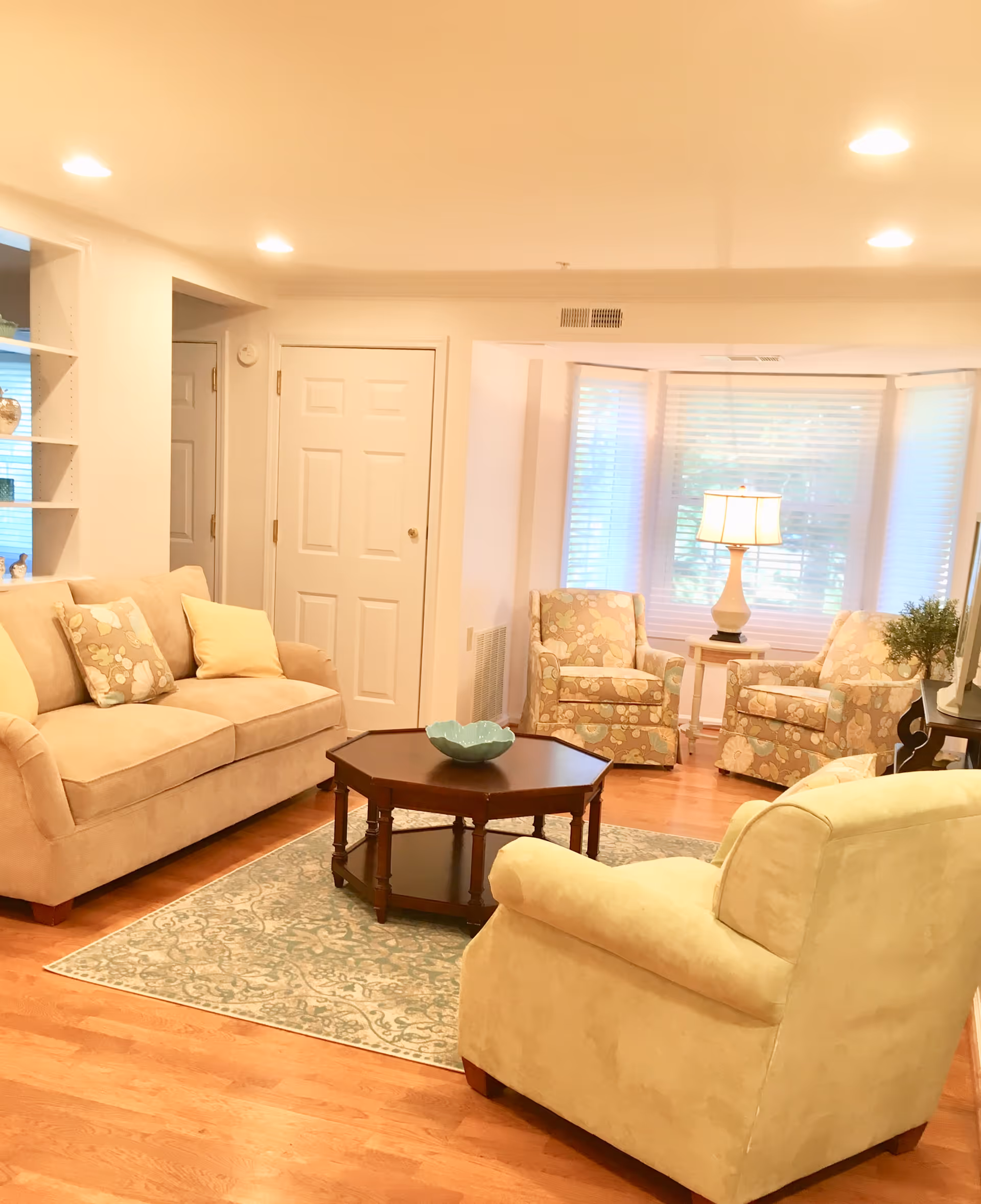 A cozy living room with a beige sofa adorned with floral and solid pillows, a matching armchair, two floral-patterned armchairs near a window with white blinds, a wooden coffee table with a decorative bowl, a side table with a lamp, and hardwood flooring with a patterned area rug.