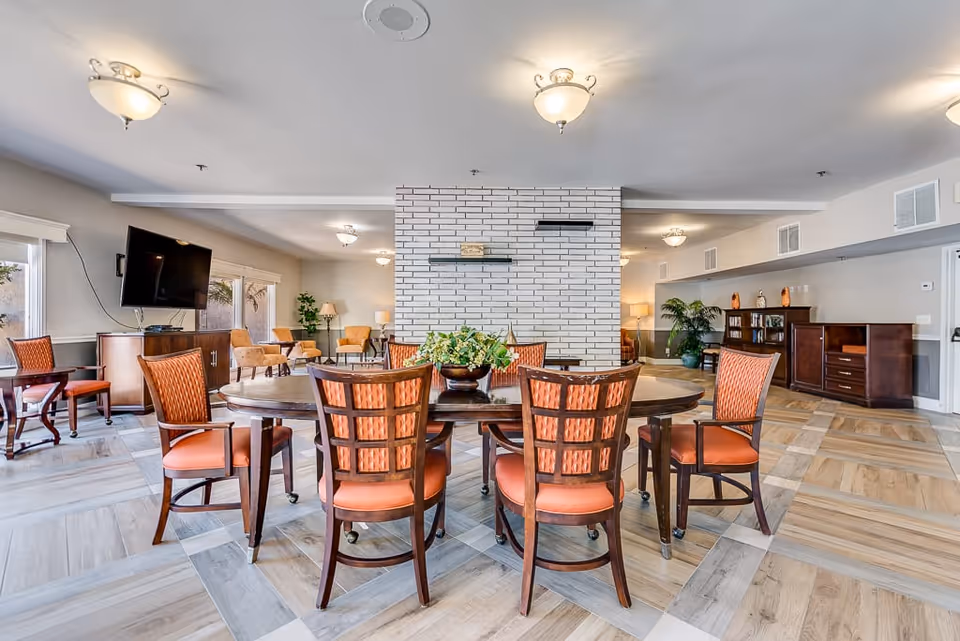 A spacious senior living common area with a round wooden table surrounded by six orange cushioned chairs. The room features a white brick fireplace in the center, wooden cabinets, a flat-screen TV mounted on the wall, and several armchairs near large windows letting in natural light. The floor has a patterned wood design, and there are ceiling lights providing warm illumination.