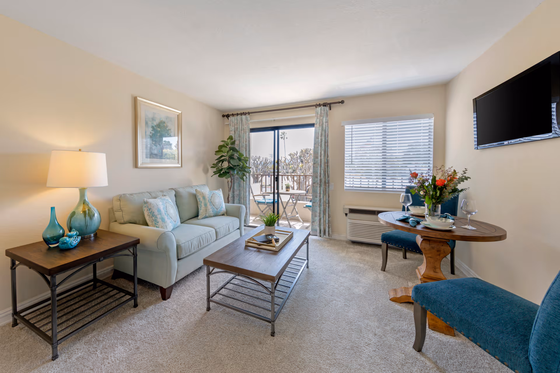 A cozy living room with a light blue sofa adorned with patterned pillows, a wooden coffee table with a small plant and tray, and a side table with a lamp and decorative glass items. There is a round wooden dining table set for two with blue upholstered chairs and a vase of flowers. A flat-screen TV is mounted on the wall, and a sliding glass door with curtains leads to a small outdoor patio with a table and chairs. A window with blinds and an air conditioning unit is also visible.