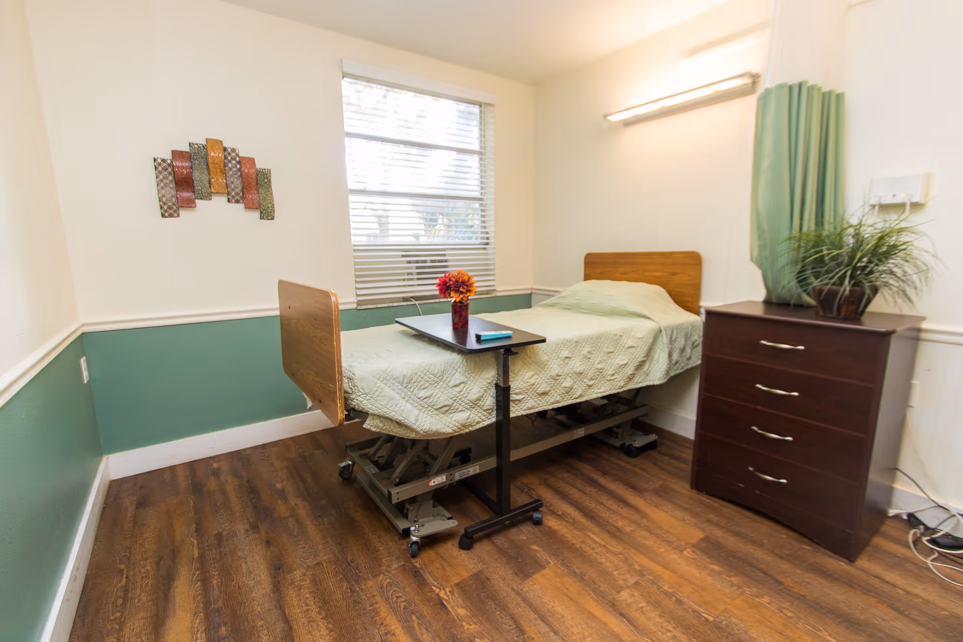 Small private room with an adjustable nursing bed covered by a green quilt, an overbed table with flowers, a wooden dresser, and a window.
