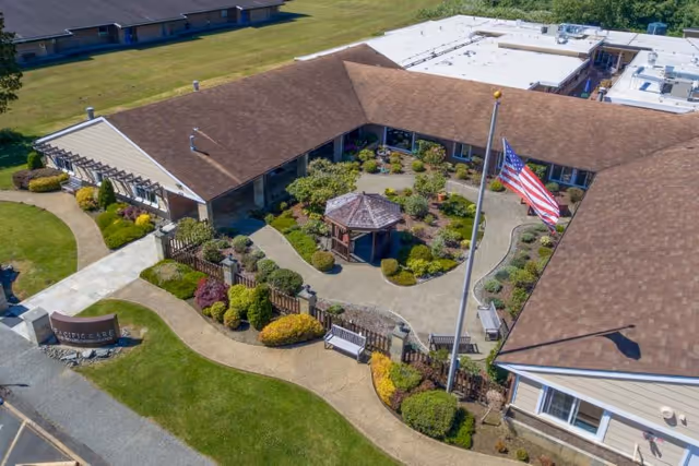 Aerial view of Pacific Care Center showing a single-story building with a brown roof surrounding a landscaped courtyard. The courtyard features a gazebo, various shrubs, and a walking path. An American flag is prominently displayed on a flagpole near the center of the courtyard. There are benches along the pathways and a sign near the entrance that reads 'Pacific Care Center'.