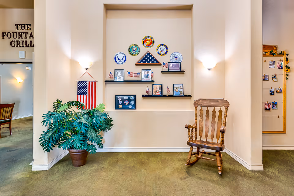 Interior wall display in a senior living facility featuring military memorabilia including an American flag folded in a triangle, military branch emblems, small American flags, and framed photographs. A wooden rocking chair is placed to the right, and a large potted plant is on the left. The entrance to a dining area labeled 'The Fountain Grill' is visible on the left side.