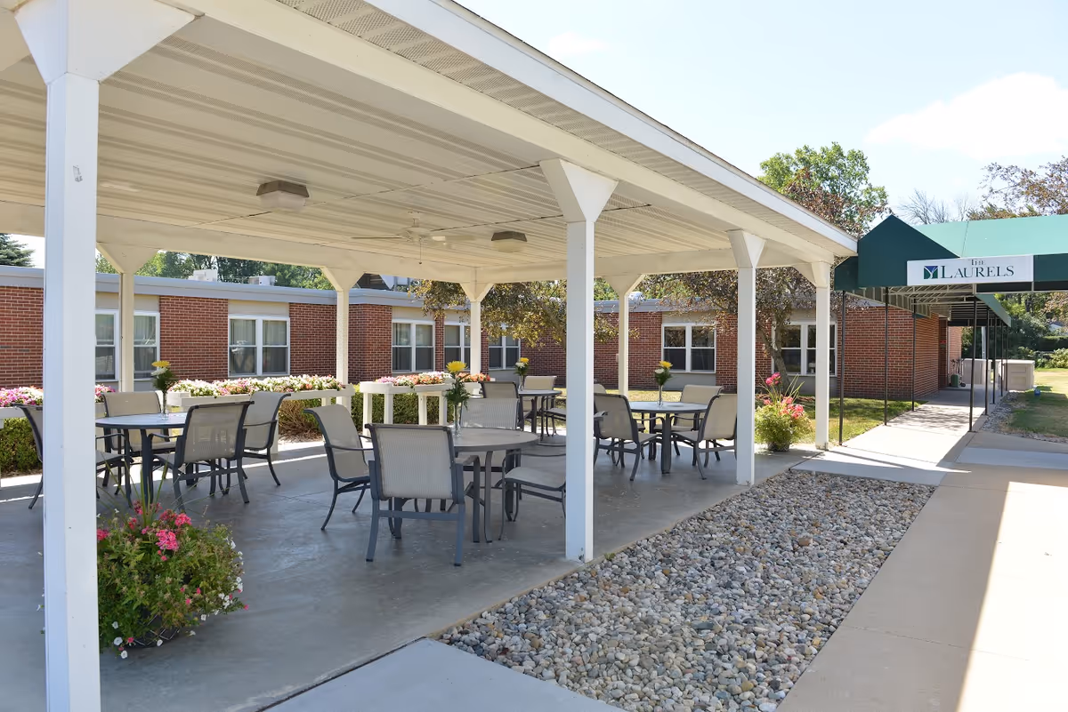 Outdoor covered patio area with several tables and chairs arranged for seating, surrounded by flower beds and greenery, adjacent to a brick building with windows. A green awning with the text 'The Laurels' is visible over a walkway leading into the building.