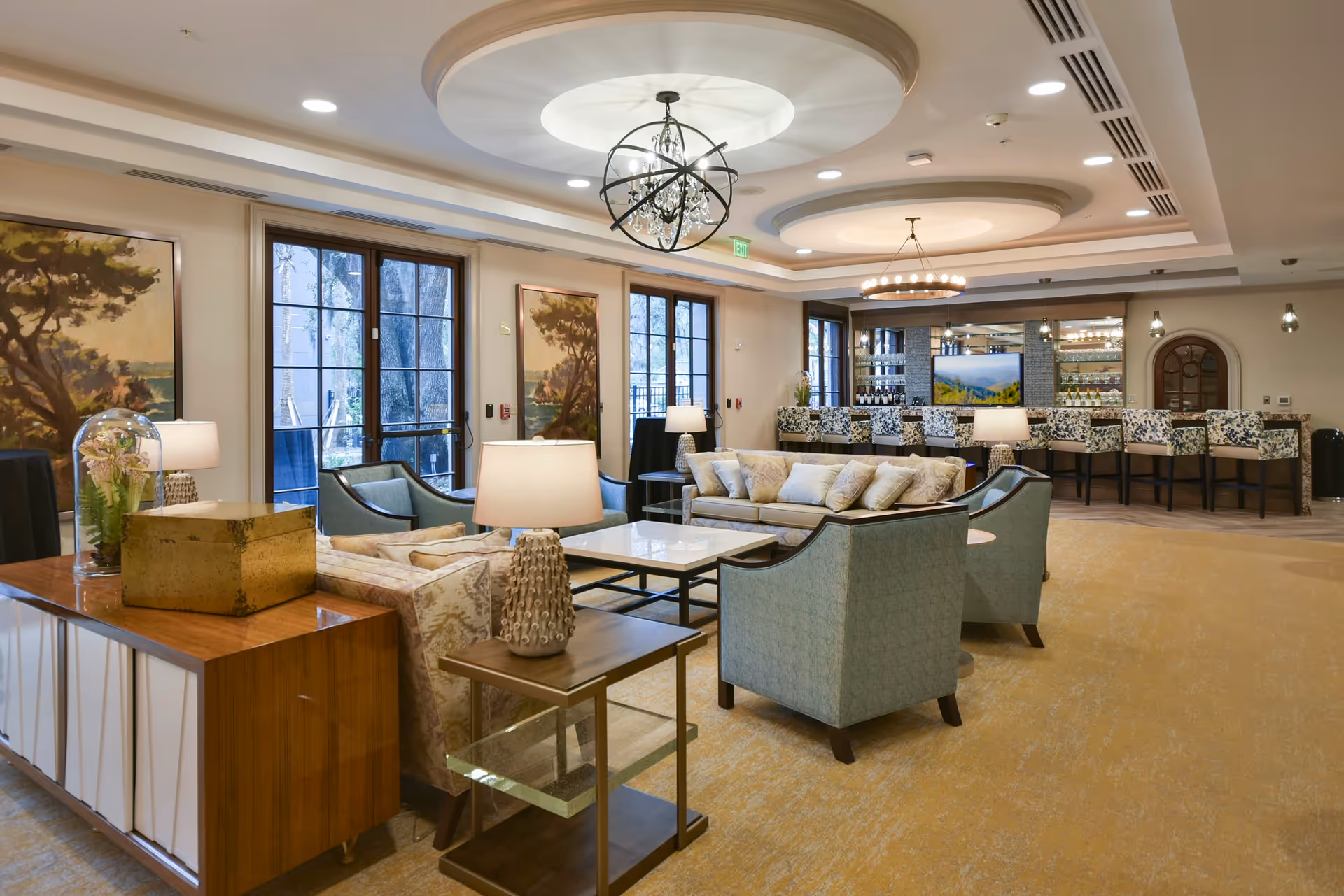 A spacious and elegantly furnished living room area in a senior living facility with comfortable sofas and armchairs arranged around a central coffee table. The room features large windows with views of trees outside, decorative paintings on the walls, and modern ceiling light fixtures. In the background, there is a bar area with high chairs and shelves stocked with bottles and glassware.