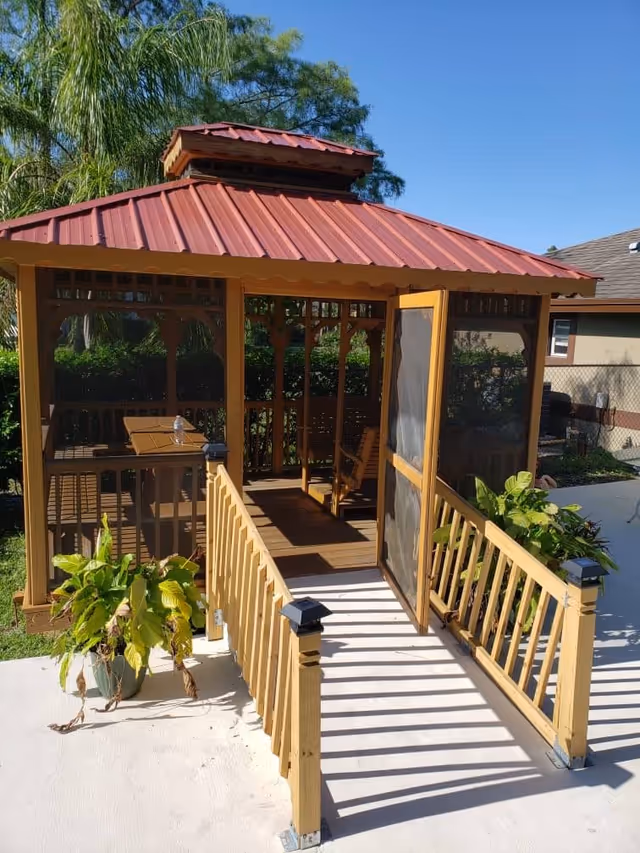 A wooden gazebo with a red metal roof and screened door, surrounded by greenery and plants, with a concrete pathway leading up to it under a clear blue sky.