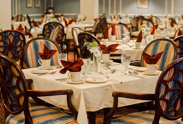 A dining room with multiple round tables covered with white tablecloths, each set with plates, cups, silverware, glasses, and red folded napkins. The chairs have colorful striped upholstery and wooden frames. A small vase with white flowers is placed on each table.