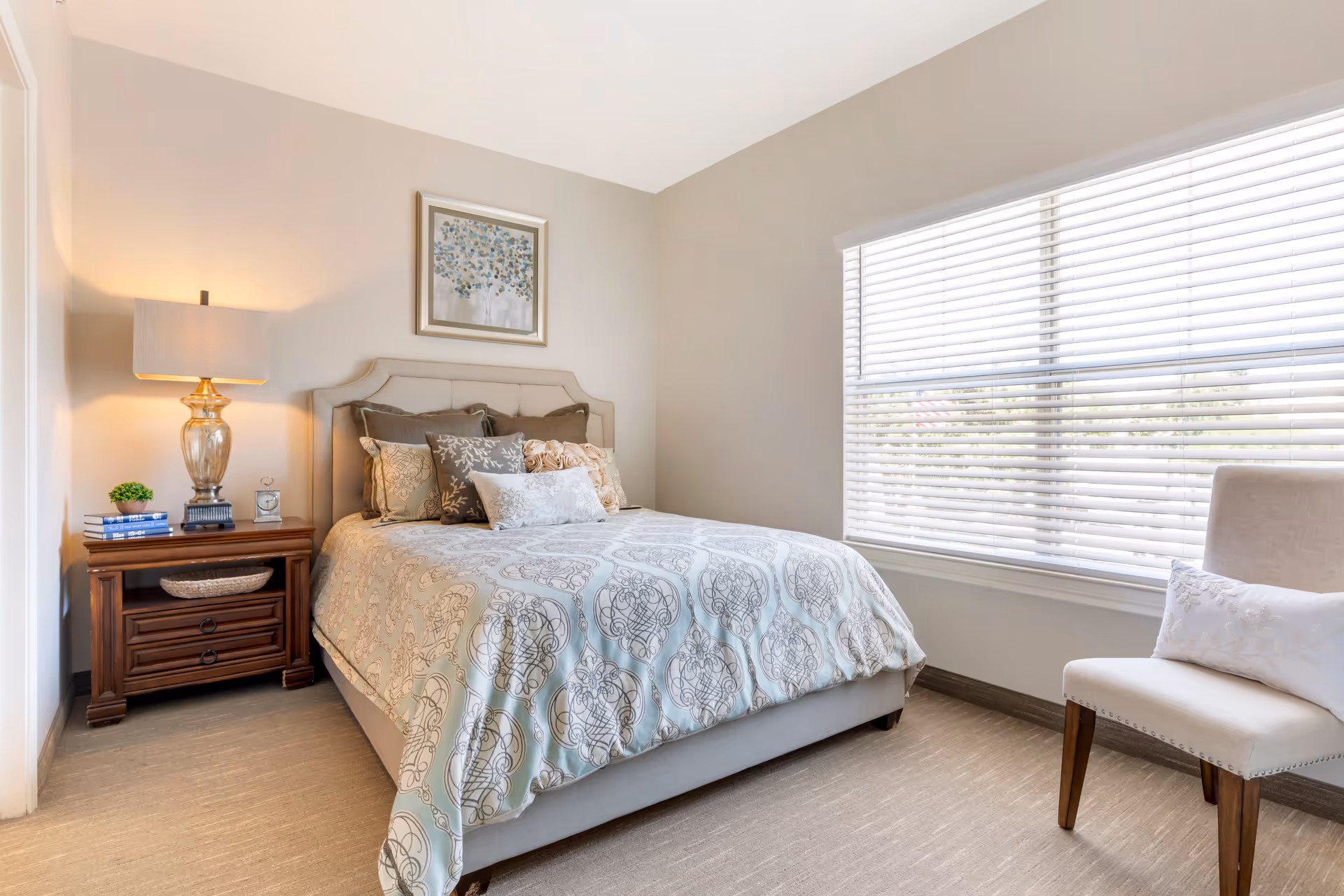 Well-lit bedroom with a neatly made bed, nightstand and lamp, large window with blinds, and a chair.