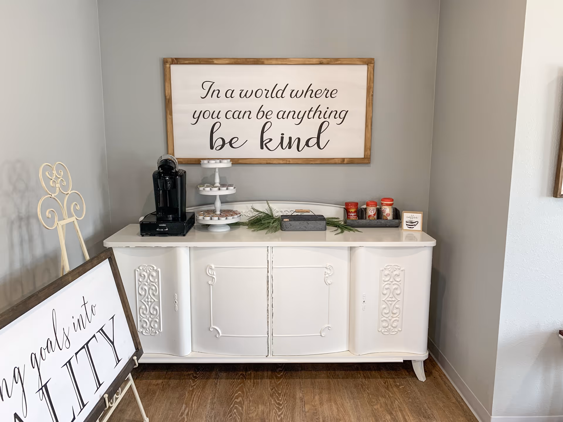 A white sideboard against a gray wall with a coffee maker, a tiered tray with small items, and coffee supplies on top. Above the sideboard is a framed sign that reads 'In a world where you can be anything be kind.' To the left, there is a decorative easel holding a sign partially visible with the words 'goals into REALITY.' The floor is wooden.