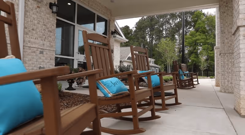 Row of wooden rocking chairs with blue cushions on a covered front porch of a brick senior living facility overlooking trees.