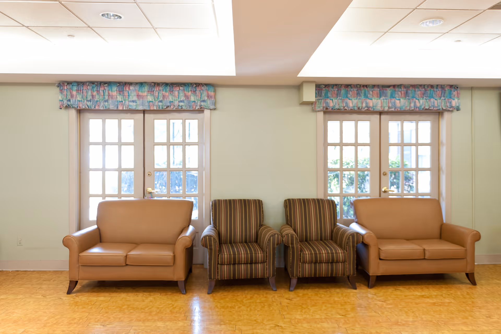 A seating area with two brown leather loveseats and two striped armchairs arranged in front of two sets of French doors with colorful valances above them. The floor is wooden and the walls are light green.