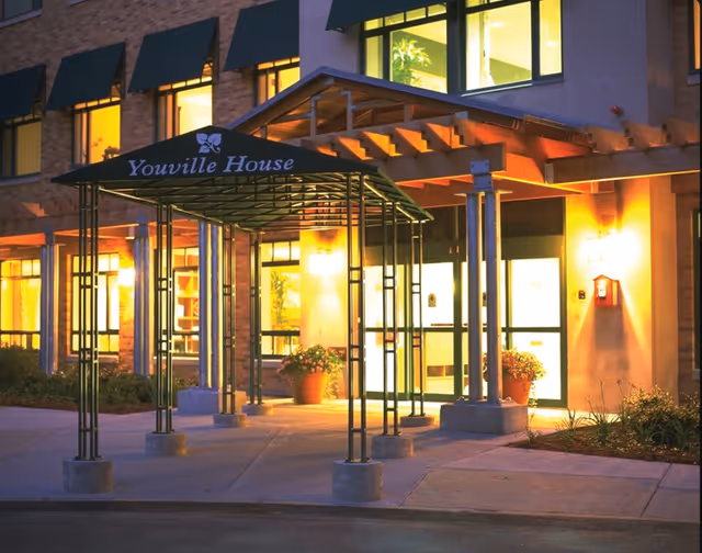 Front entrance of Youville House assisted living featuring a covered metal canopy, potted plants, and illuminated glass doors at dusk.
