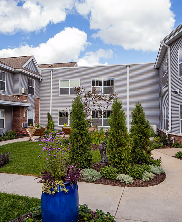 Outdoor courtyard area of a senior living facility with a well-maintained garden featuring various shrubs, a blue flower pot with purple flowers, a small statue, and pathways surrounded by green grass and landscaping. The building exterior is visible with gray siding and multiple windows under a partly cloudy sky.