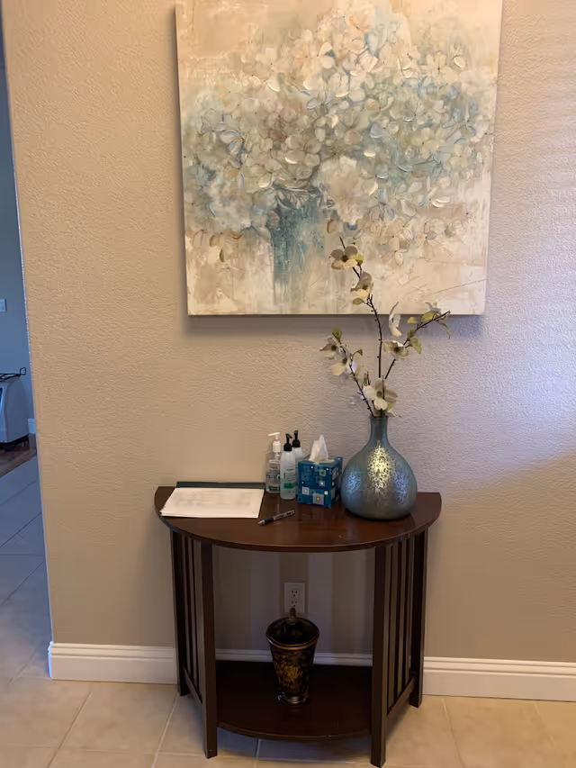 A small wooden half-moon table against a beige wall with a large floral painting hanging above it. On the table are a blue vase with white flowers, a tissue box, hand sanitizer, lotion, a pen, and a paper. Below the table is a decorative container. The floor is tiled and there is a doorway visible to the left.